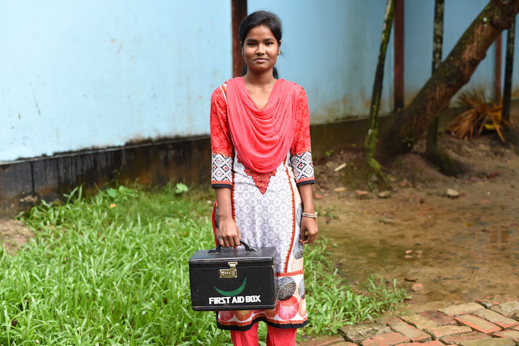 Teenage girl wearing pink and whte is holding a first aid box and standing in front of her school.